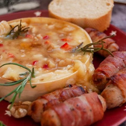 A baked camembert with red chillies and rosemary sprigs on a red plate with sausages wrapped in bacon
