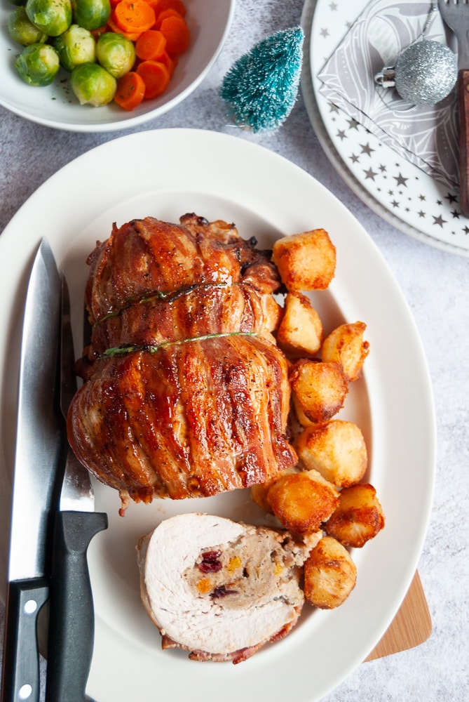 A cooked rolled and stuffed turkey breast joint tied up with string on a white plate with a carving fork and knife, serving plates and a bowl of carrots and Brussels sprouts.