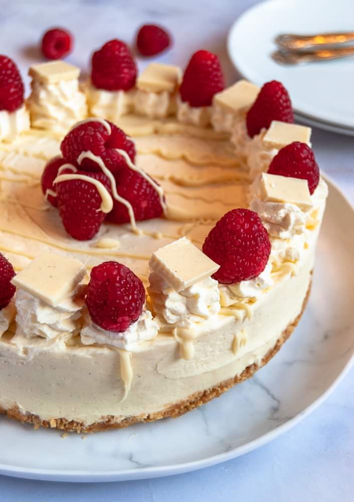 A white chocolate and raspberry cheesecake on a white plate decorated with fresh raspberries, whipped cream and melted white chocolate