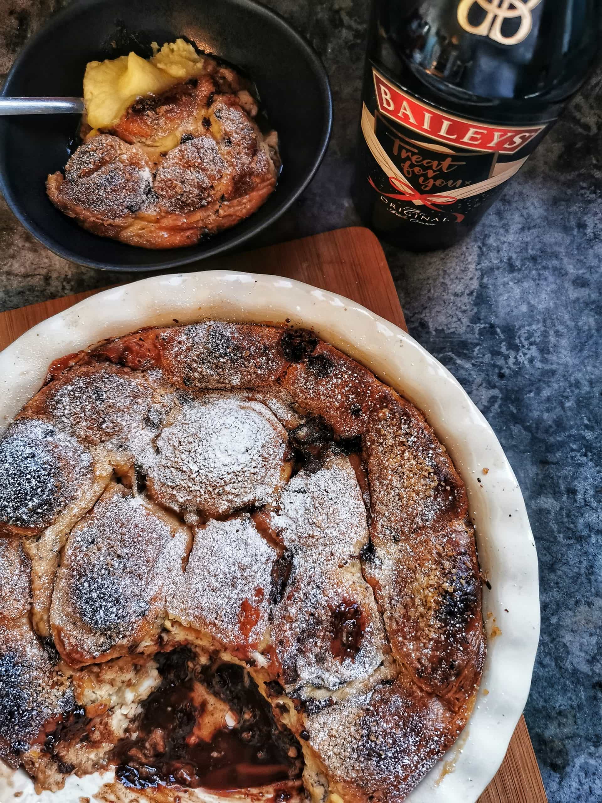 A chocolate baileys brioche pudding in a round dish. A bottle of baileys and a small bowl containing the pudding with cream can also be seen 