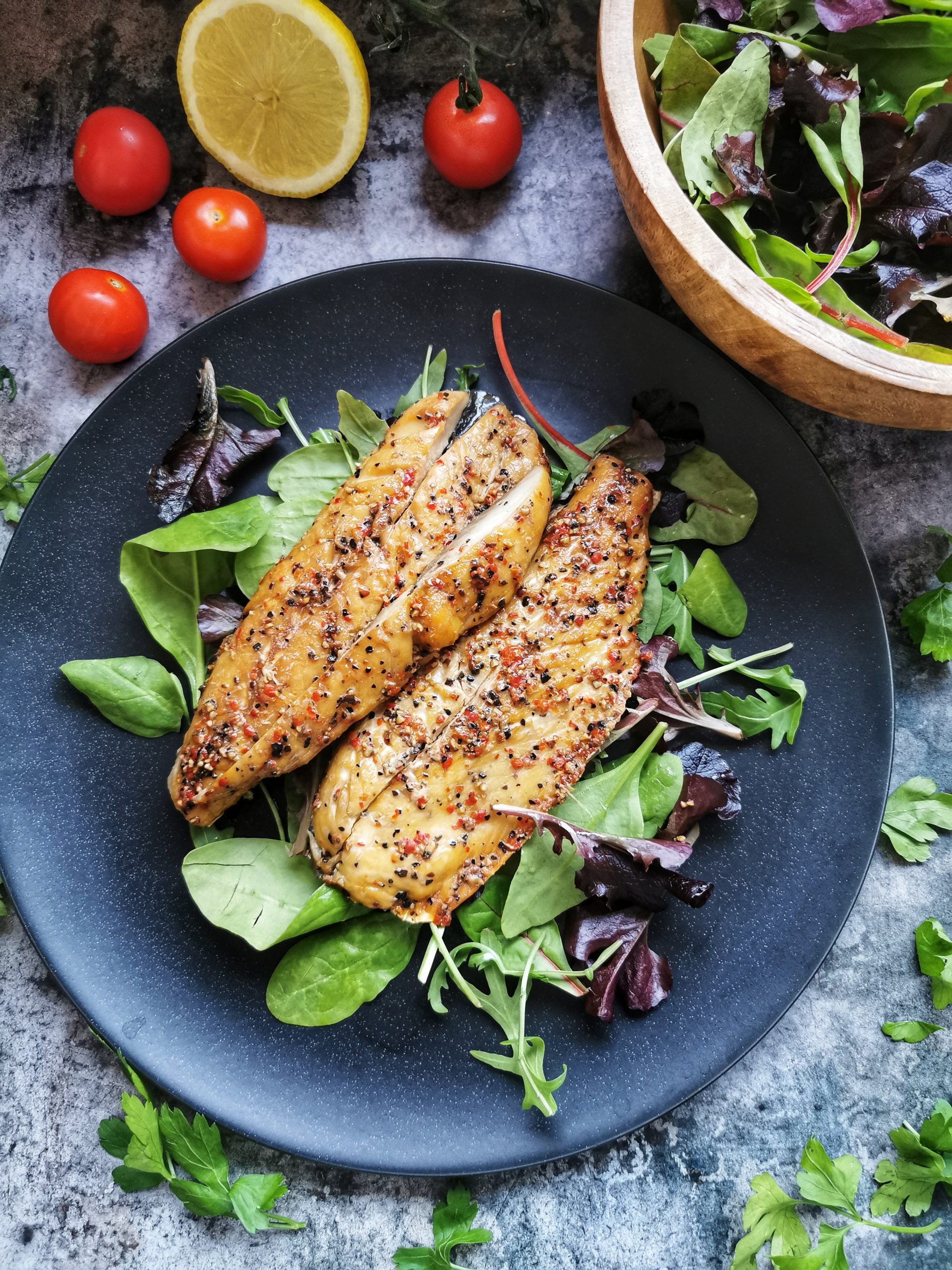 Two peppered mackerel fillets and salad on a black plate, vine tomatoes, half a lemon and a bowl of salad leaves.