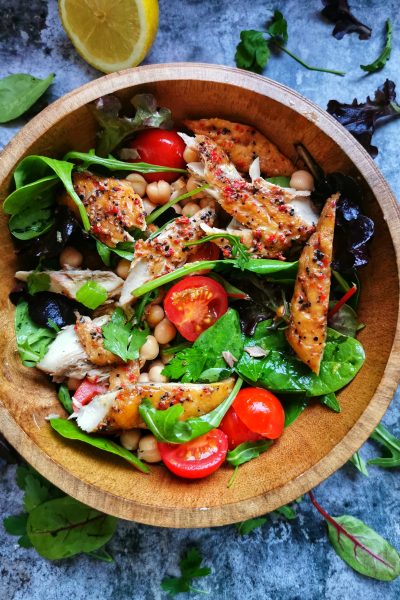 A wooden bowl of salad leaves, smoked mackerel, tomatoes and chickpeas on a grey background