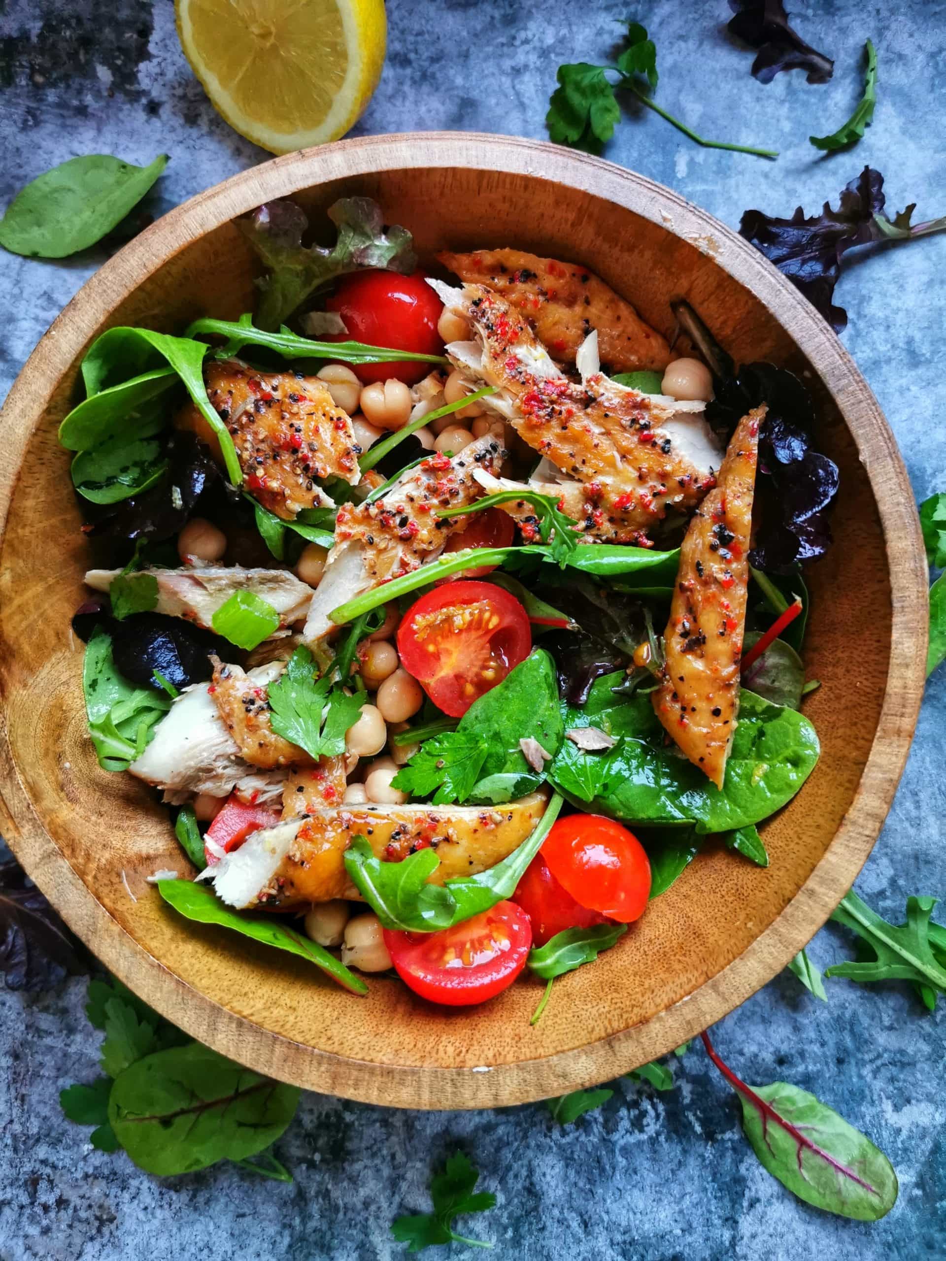 A wooden bowl of salad leaves, smoked mackerel, tomatoes and chickpeas on a grey background