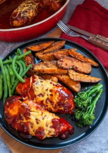 A black plate of 2 chicken breasts covered in BBQ sauce and melted cheese with sweet potato wedges, green breans and tenderstem broccoli.