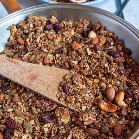 A pan of granola with dried cranberries and nuts. A wooden spoon rests against the pan.