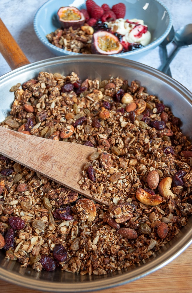 A pan of granola with dried cranberries and nuts. A wooden spoon rests against the pan.  A blue bowl of granola with yoghurt, raspberries and passionfruit sits in the background.