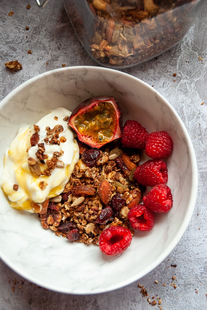 A white bowl filled with granola, yoghurt, passion fruit and raspberries on a grey background 