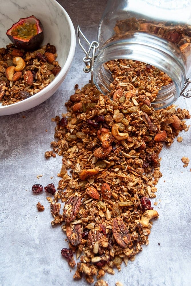 A large jar of granola with nuts, seeds and dried cranberries and sultanas spilling out onto a work surface.