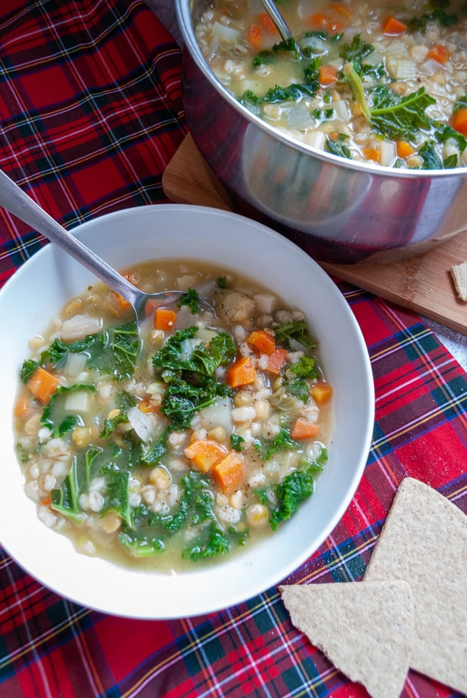 A bowl of vegetable soup with a silver spoon and oatcakes on the side