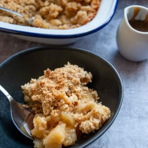 A bowl of apple crumble with a small white jug of toffee sauce on the side. A dish of the crumble can be seen in the background.