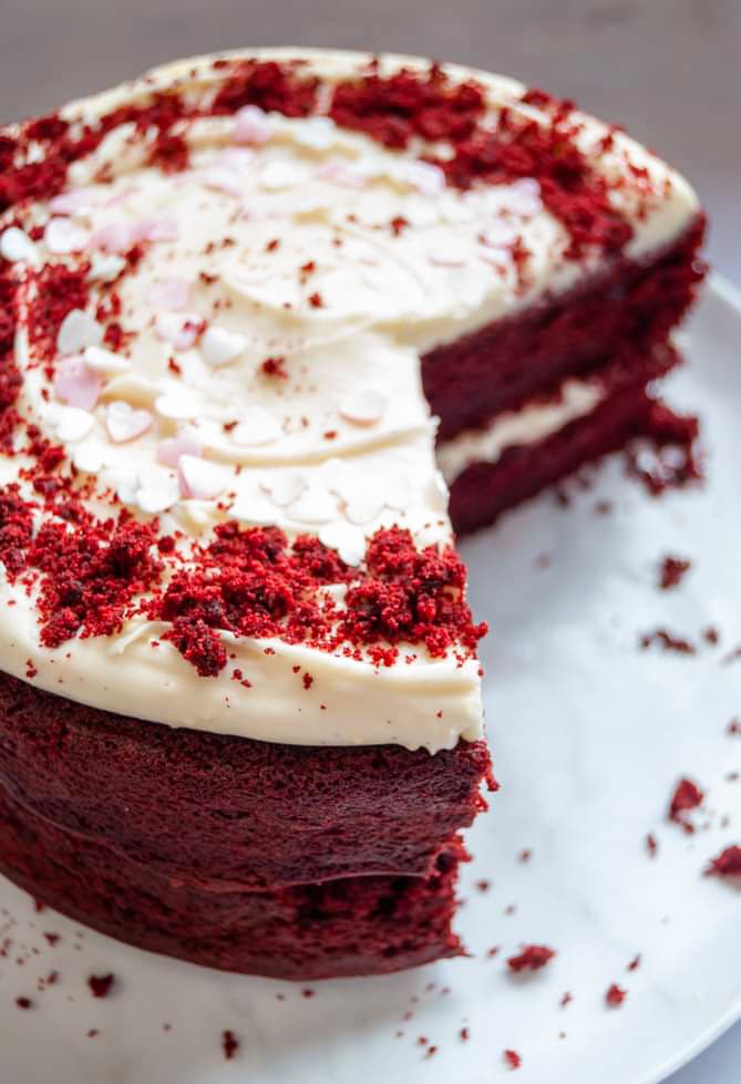 A close up picture of a red velvet cake with cream cheese frosting and pink heart sprinkles on a white Plate 