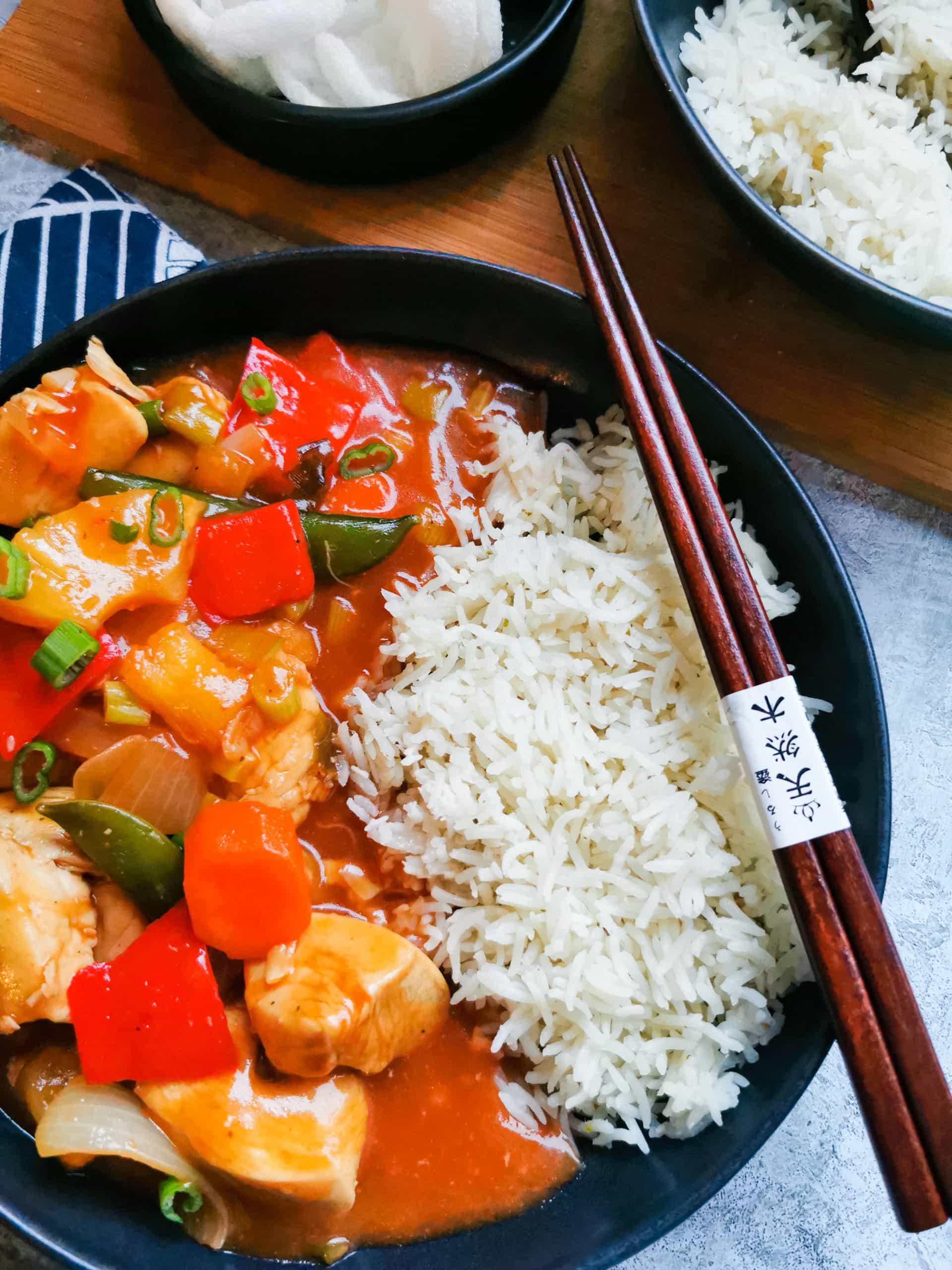 A flat lay photo of a black plate of sweet and sour chicken with rice and a pair of wooden chopsticks resting against the plate.