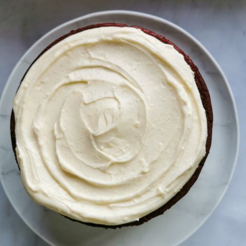 A red cake covered with cream cheese frosting on a white plate.