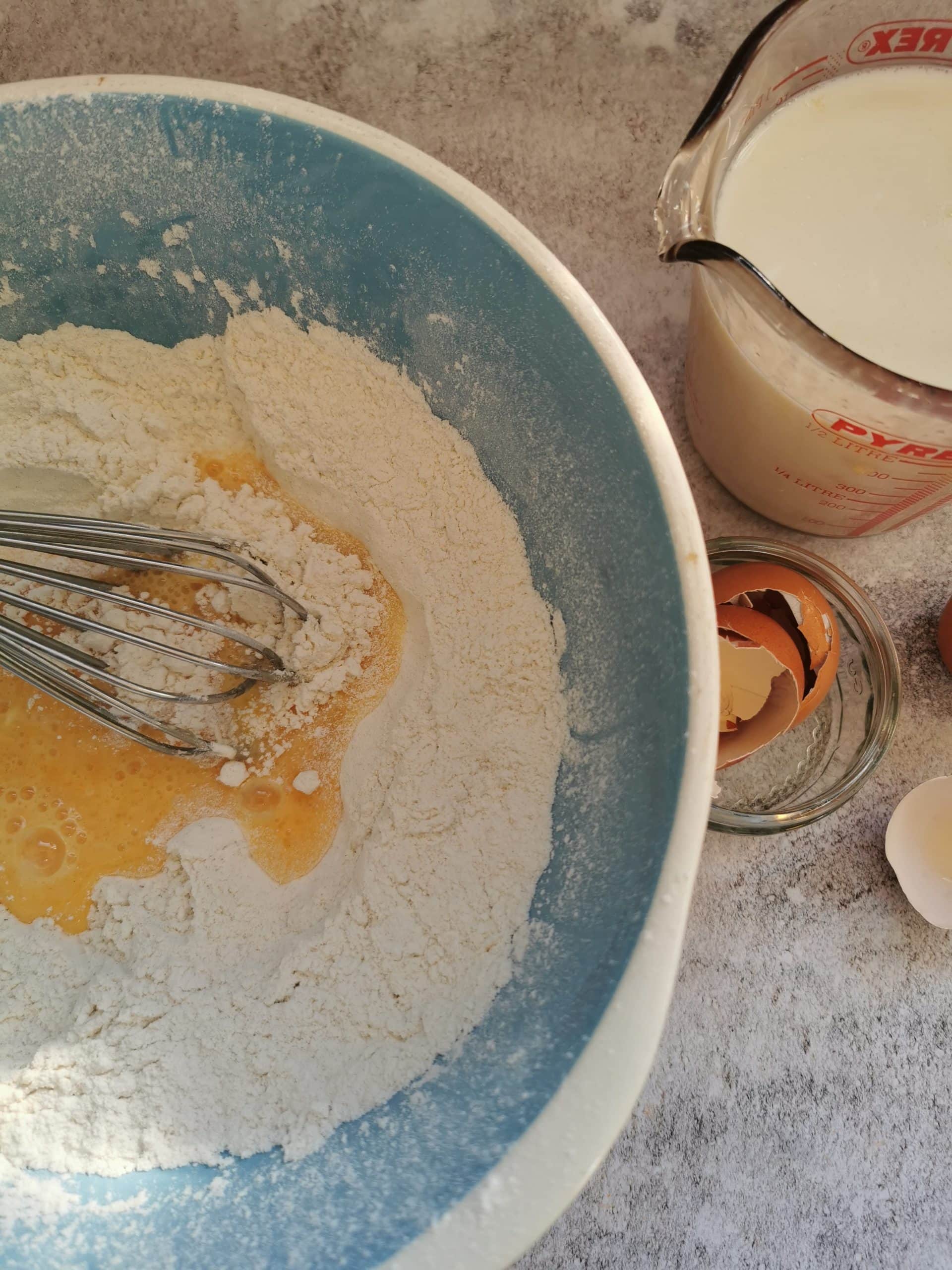 A large blue and white mixing bowl with flour, eggs and a whisk
