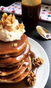 A close up pancake stack covered in toffee sauce and topped with whipped cream and chopped walnuts