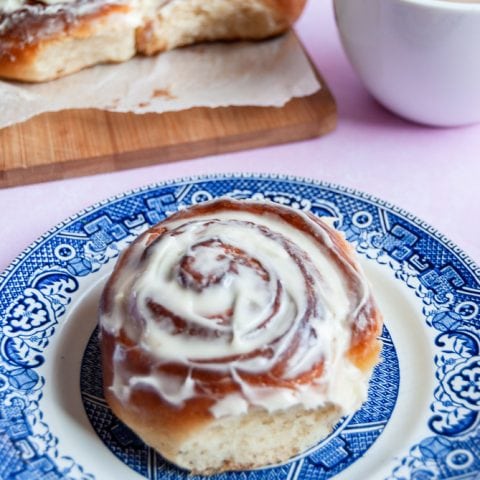 A frosted cinnamon roll on a blue willow pattern plate. A white cup of coffee and more cinnamon rolls on a wooden board