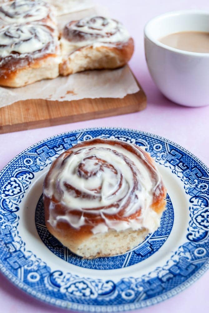 A frosted cinnamon roll on a blue willow pattern plate. A white cup of coffee and more cinnamon rolls on a wooden board