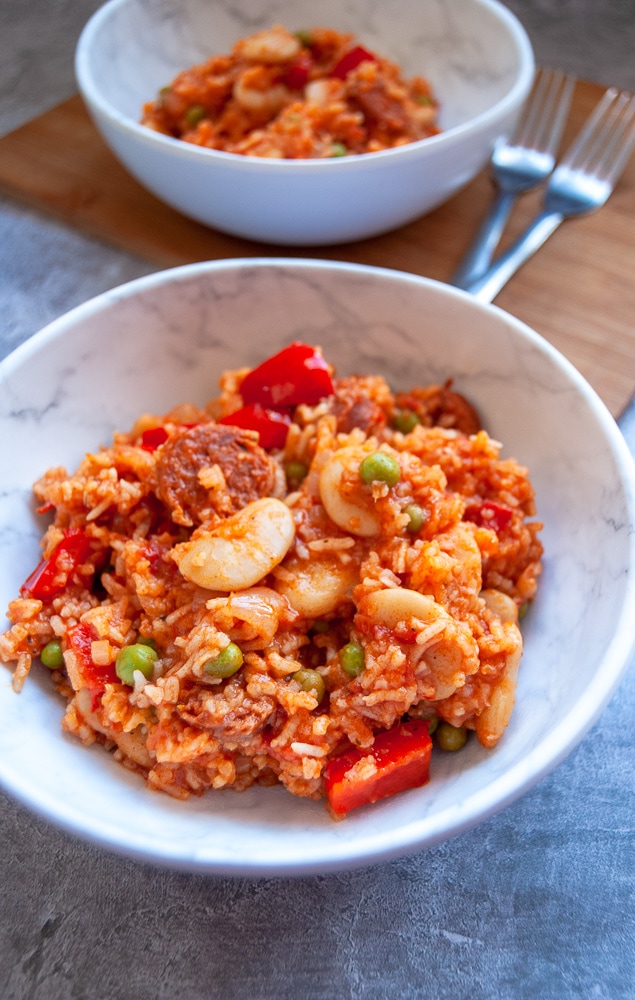 A white bowl of rice with red peppers, chorizo and peas