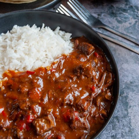 A black stoneware bowl filled with beef curry and basmati rice. A plate of poppadoms and a bowl of mango chutney in the background