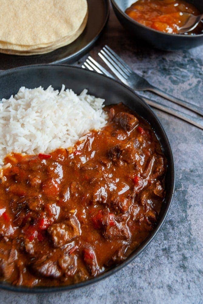 A black stoneware bowl filled with beef curry and basmati rice. A plate of poppadoms and a bowl of mango chutney in the background