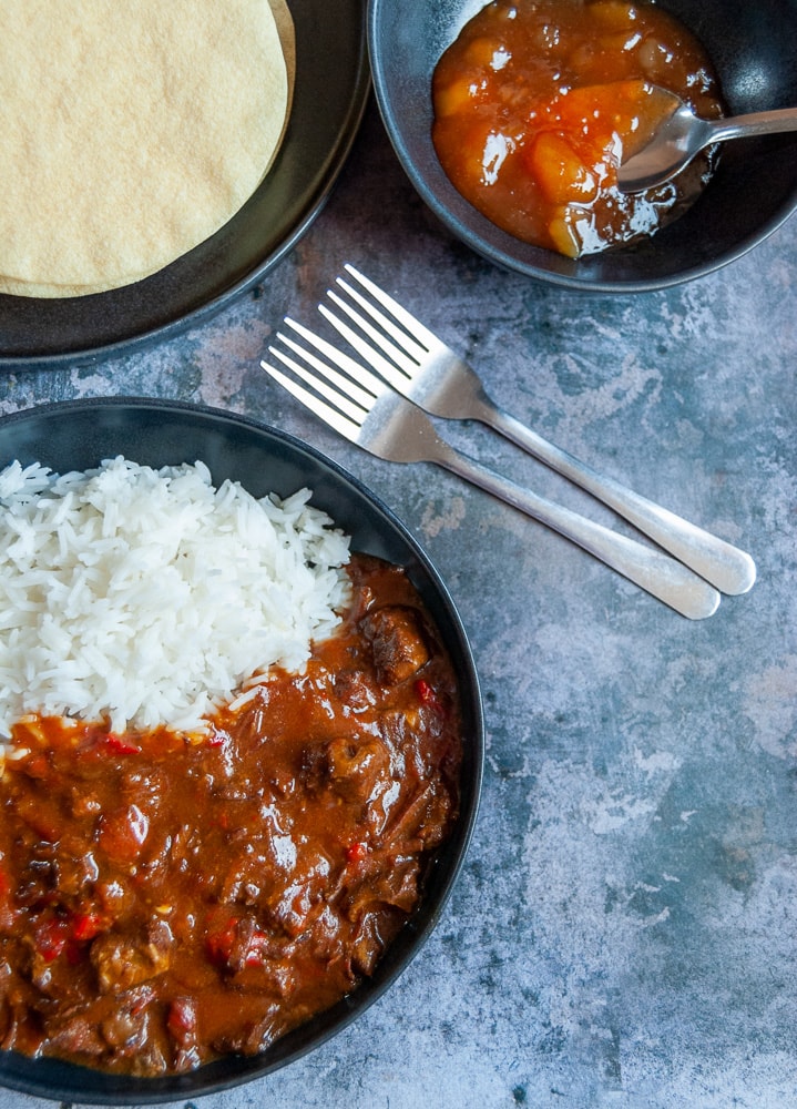 A flat lay photo of a bowl of beef curry with red chillies and basmati rice, a bowl of mango chutney and a plate of poppadoms