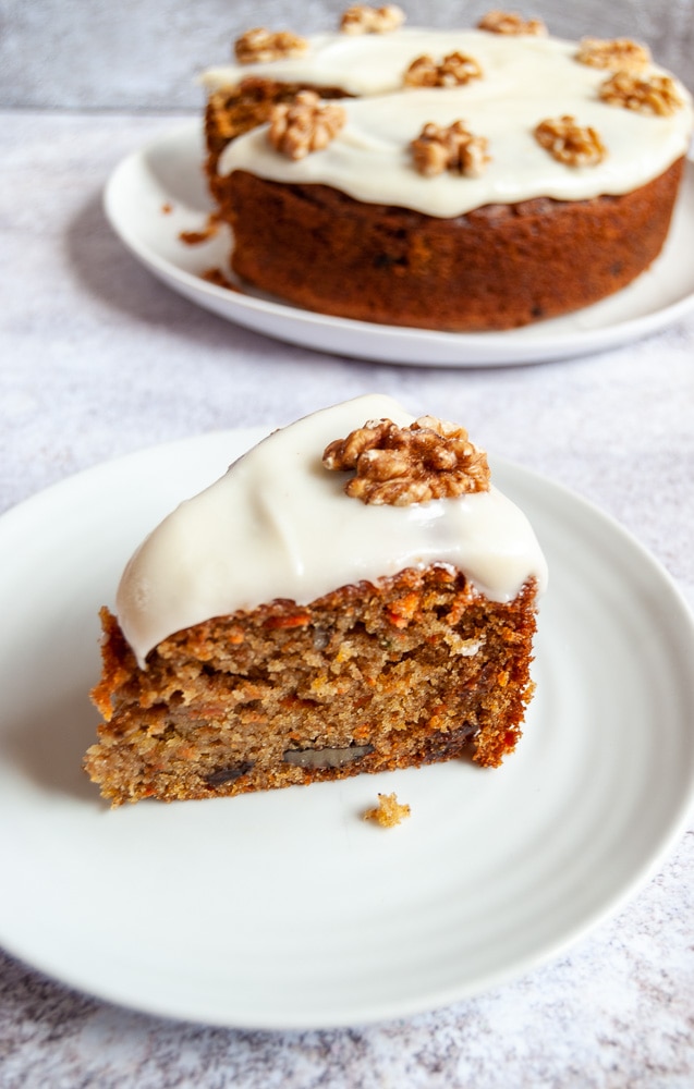 A slice of carrot cake topped with cream cheese frosting and a whole walnut on a white plate. A large carrot cake can be seen in the background.