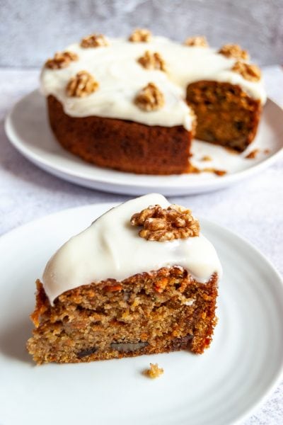 A slice of carrot cake topped with cream cheese icing and a walnut on a white plate.
