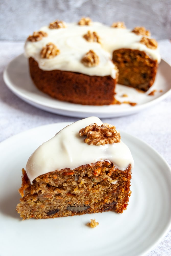 A slice of carrot cake topped with cream cheese icing and a walnut on a white plate.