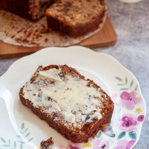 A slice of buttered fruit loaf on a pink and yellow floral cake. A fruit loaf can be seen on a wooden board in the background.