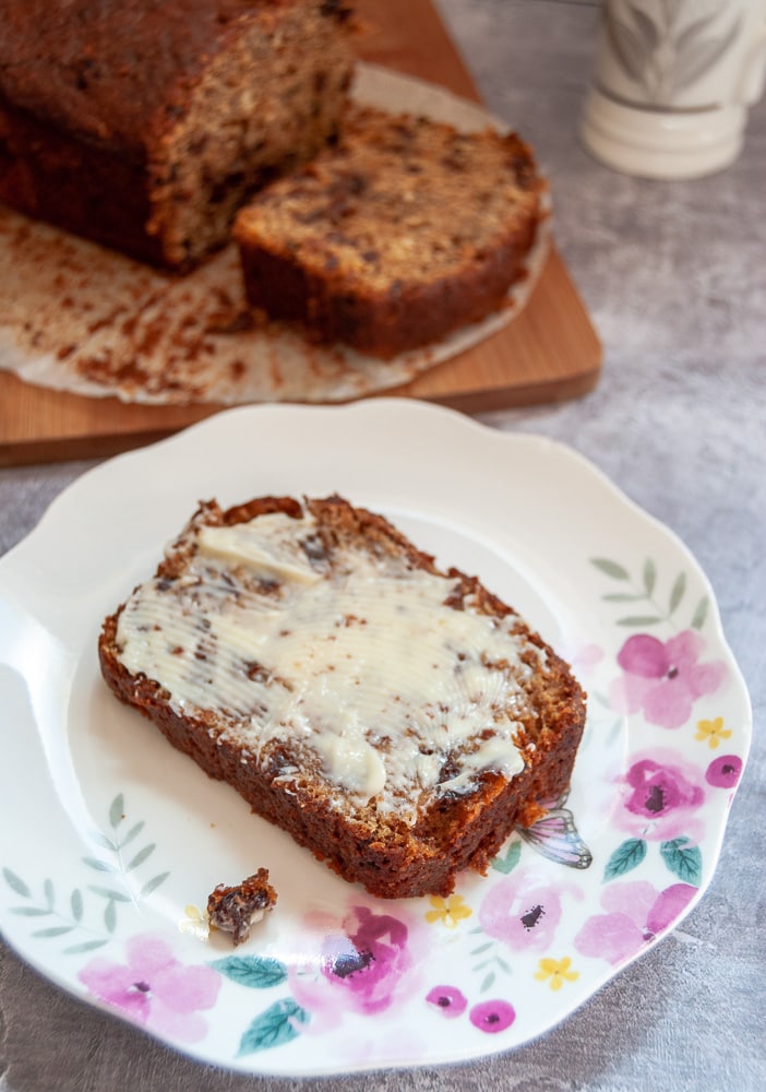 A slice of buttered fruit loaf on a pink and yellow floral cake. A fruit loaf can be seen on a wooden board in the background.