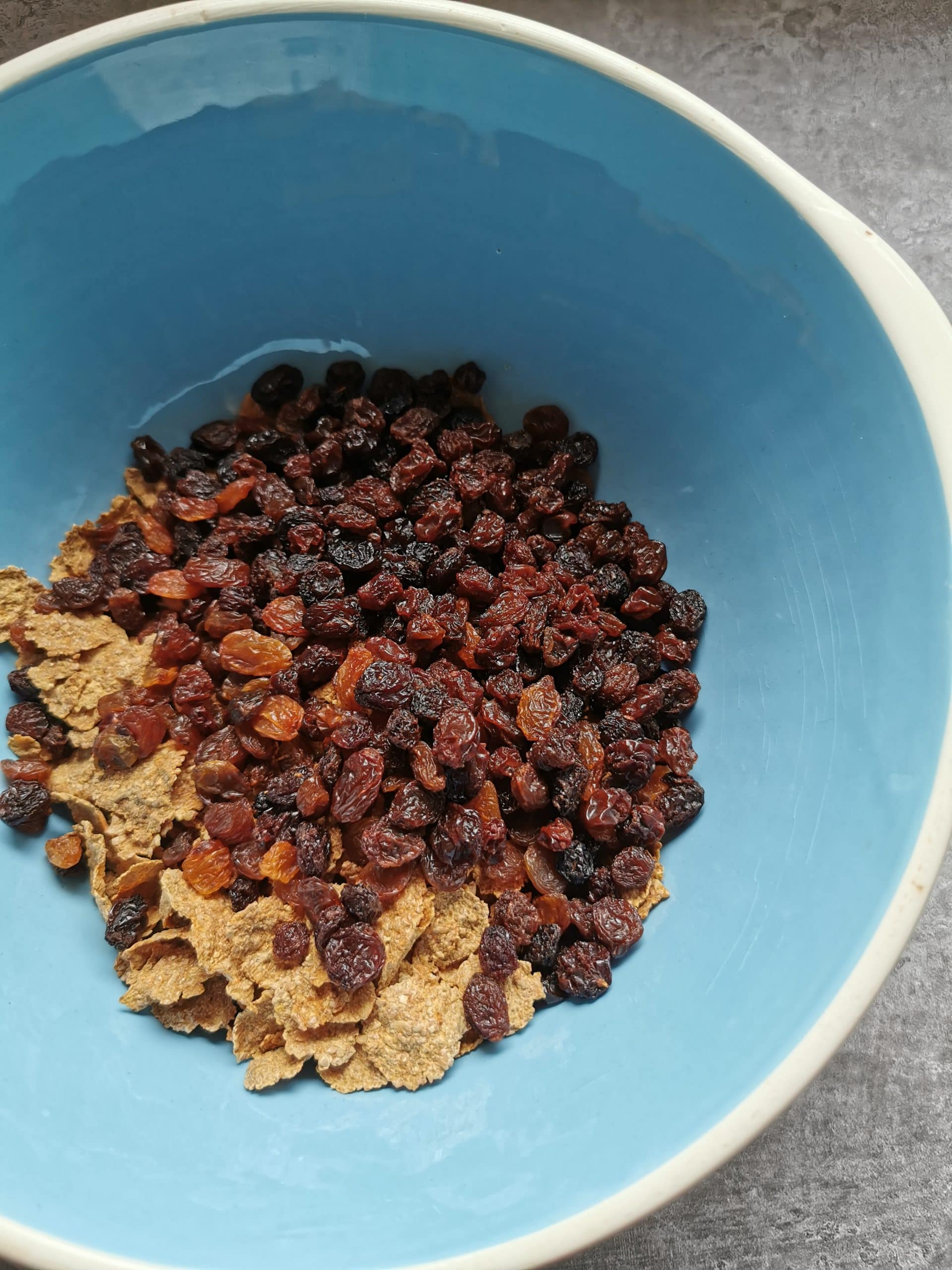 A blue and white bowl of bran flakes, sugar and dried fruit.