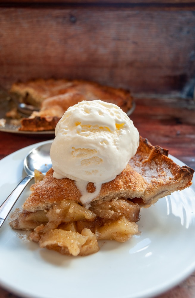 A slice of apple pie on a white plate topped with a scoop of vanilla ice cream