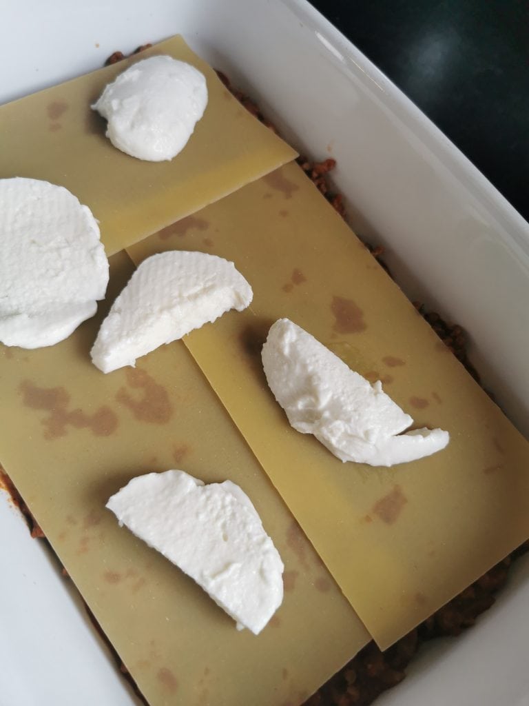A lasagne being layered with mozzarella cheese in a white dish 