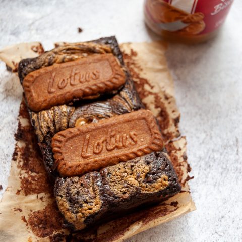 Two chocolate brownies topped with Biscoff spread and two Biscoff cookies and a jar of biscoff spread