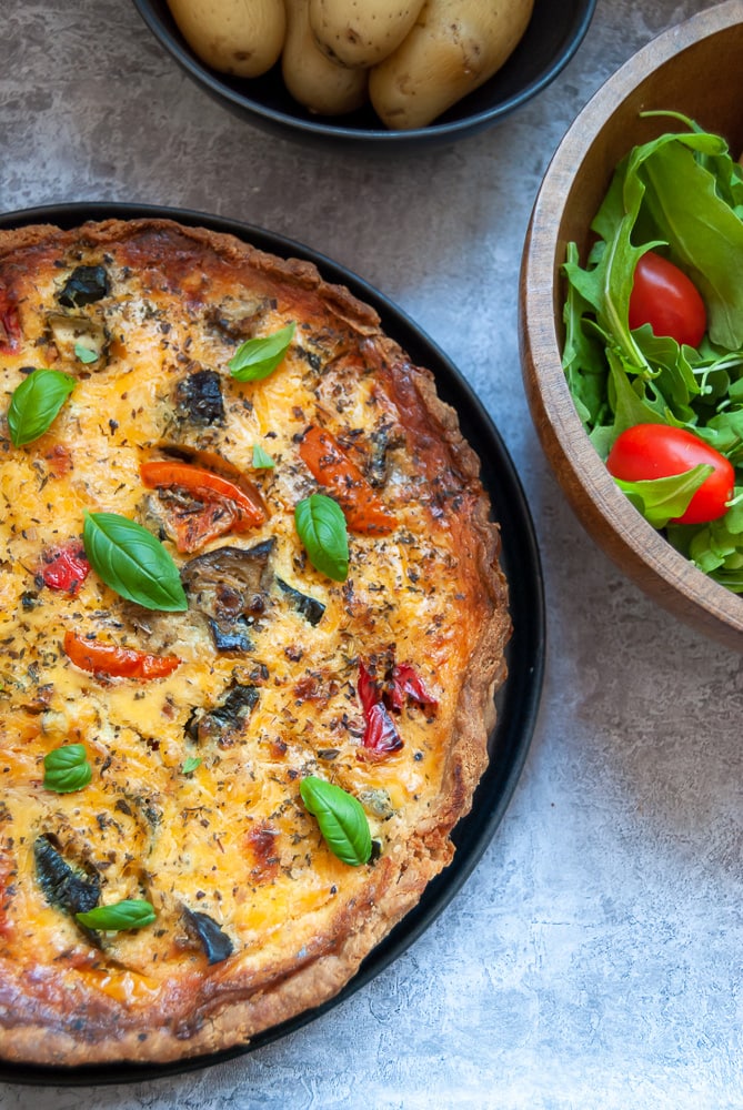 A quiche filled with aubergine, tomatoes, peppers and scattered with fresh basil leaves on a black plate, a wooden salad bowl filled with green salad leaves and tomatoes and a bowl of potatoes.