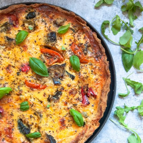 A quiche filled with aubergine, tomatoes, bell peppers and scattered with fresh basil leaves on a grey-blue backdrop. Fresh herbs are scattered around the quiche.
