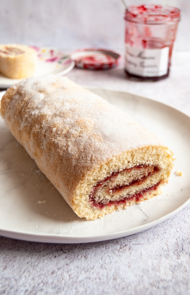 A swiss roll filled with jam and sprinkled with sugar on a white plate, a small plate with a piece of cake and a jar of raspberry jam with a spoon