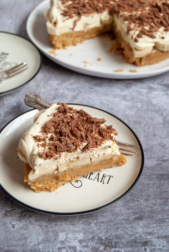 a slice of banoffee pie with biscuit base, caramel, bananas, whipped cream and grated chocolate on a white and black plate. The large pie can be partially seen in the background.