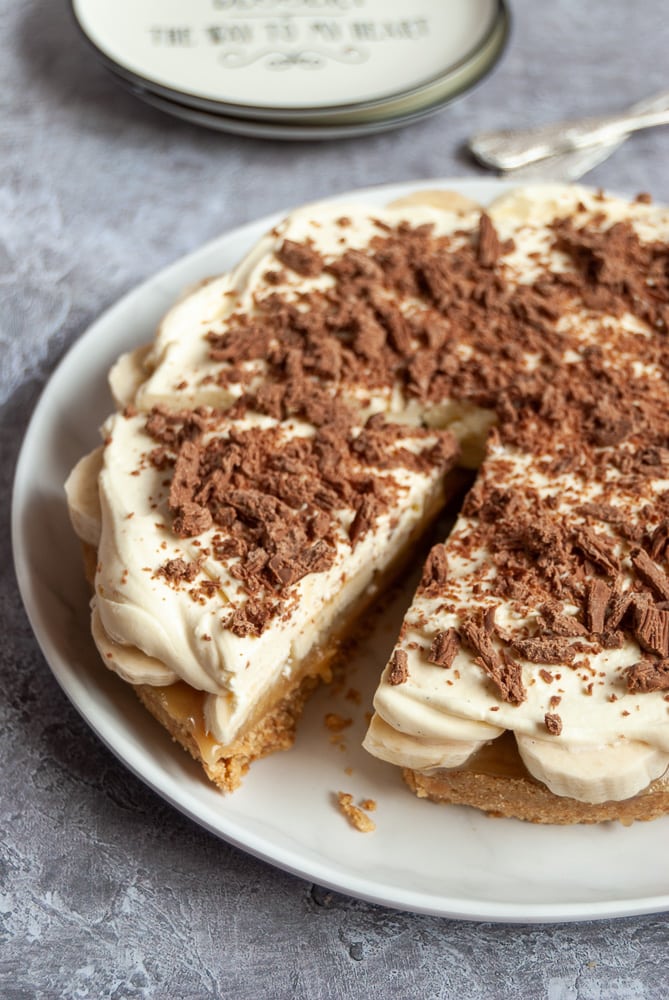 A banoffee pie covered in softly whipped cream and grated chocolate with a slice cut on a white plate.