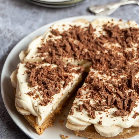 A banoffee pie covered in softly whipped cream and grated chocolate with a slice cut on a white plate.