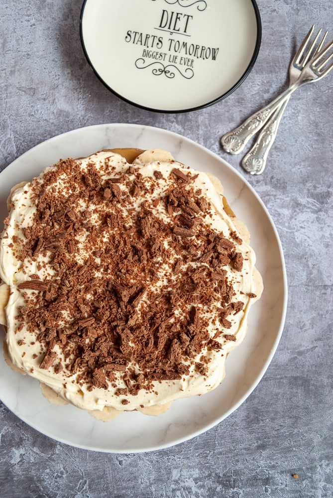 a flat lay photo of a banana cream pie covered in grated chocolate on a white plate, two silver cake forks and a black and white serving plate.