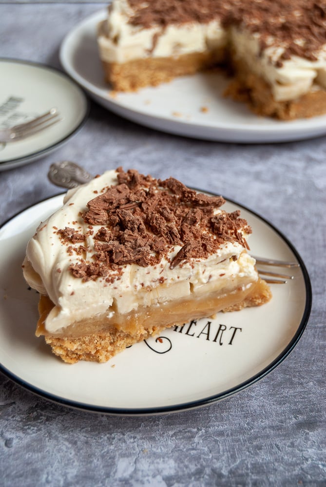 a slice of banoffee pie covered in whipped cream and grated chocolate on a white and black plate. The large cut pie can be partially seen in the background.