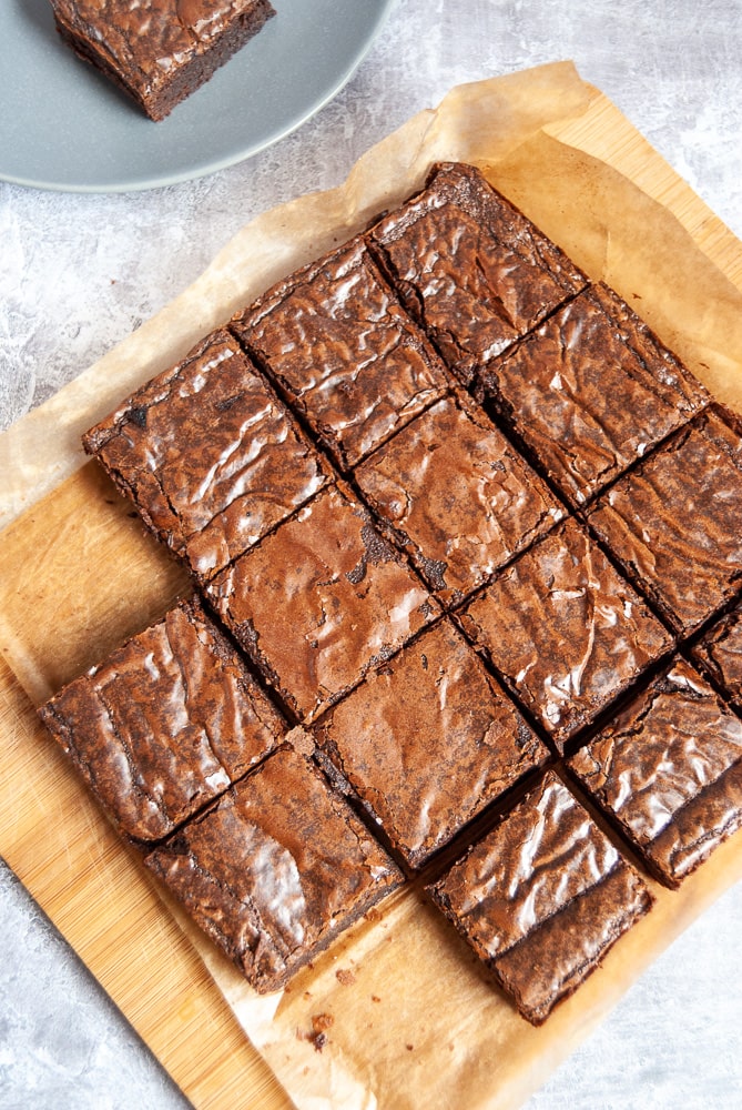 A batch of chocolate brownies on a wooden board. A grey plate with a brownie on it can be partially seen.