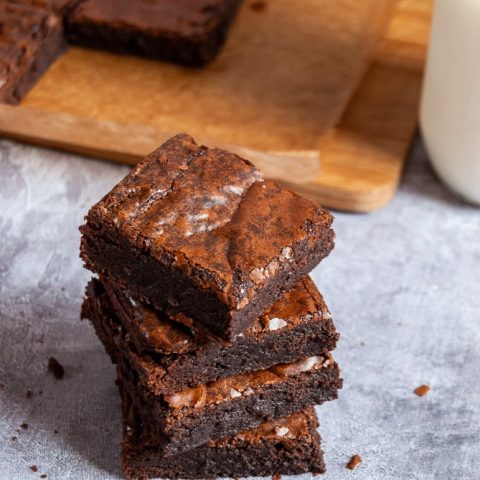 A stack of brownies and more brownies on a wooden board. A glass bottle of milk can be partially seen.