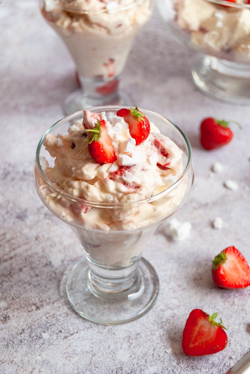 A serving glass of strawberry Eton mess, broken meringues and halved strawberries on a grey background