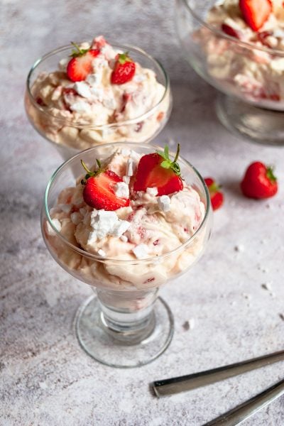 Two dessert glasses filled with whipped cream, strawberries and crushed meringue on a grey background. Two dessert spoons can be partially seen
