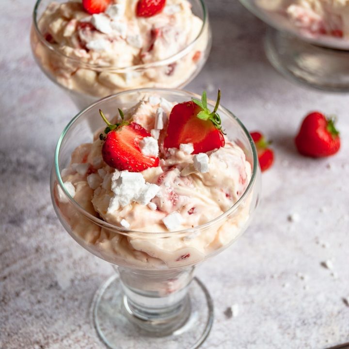 Two dessert glasses filled with whipped cream, strawberries and crushed meringue on a grey background. Two dessert spoons can be partially seen