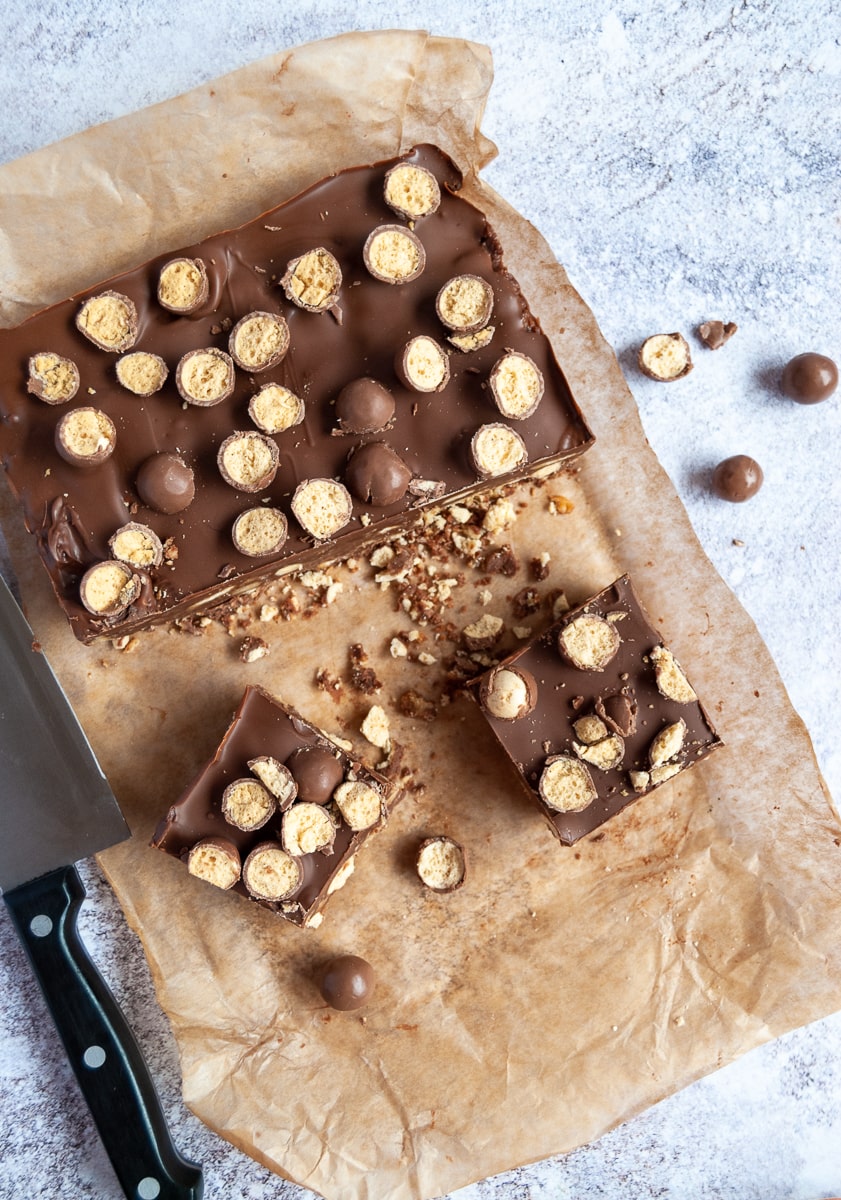 A partially cut Malteser chocolate slice on a crumpled square of baking paper and a large black handled knife