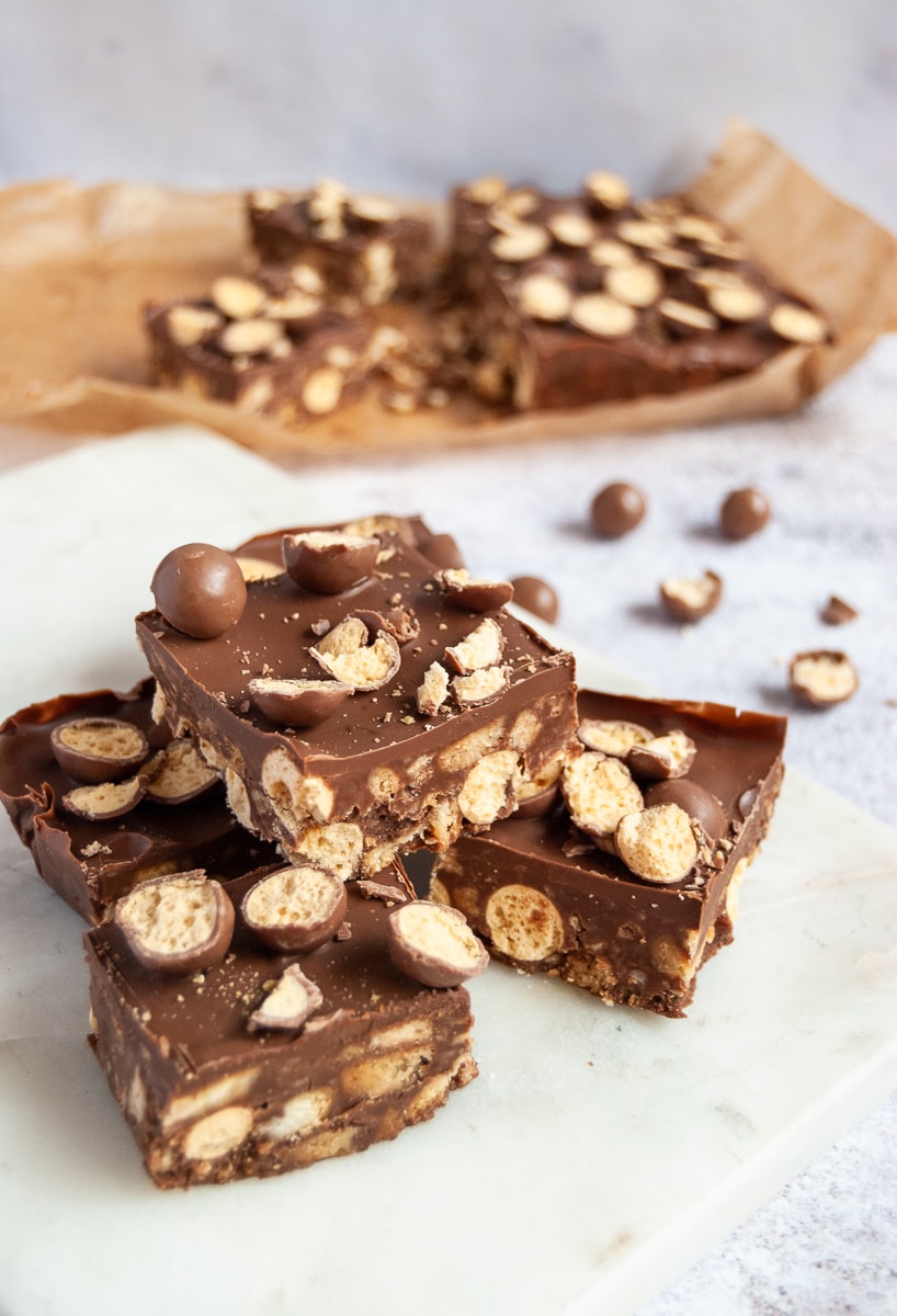 Square of Malteser traybake piled on top of each other on a white marble board