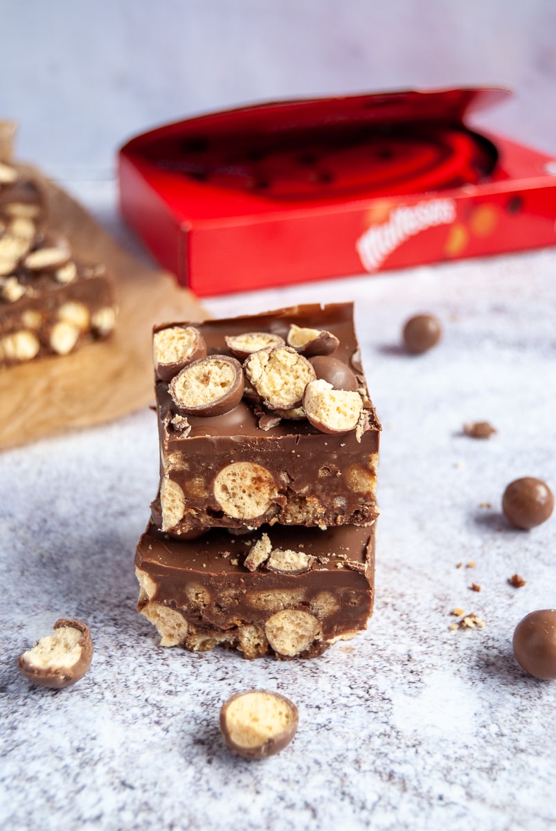 two pieces of malteser slice on top of each other on a grey-blue background with a box of Maltesers in the background